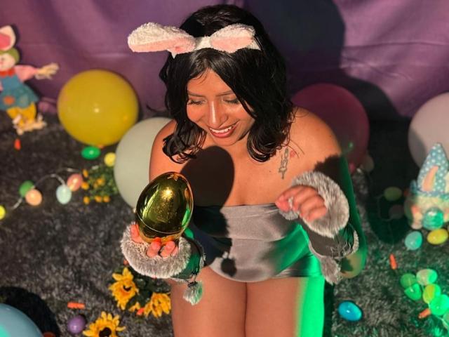 ZarayWhitMoreLove with dark hair, pink bunny ears, gray strapless bodysuit, holding shiny golden egg, smiling, fluffy gray cuffs, colorful balloons, and green light on gray carpet.