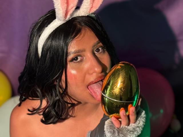 ZarayWhitMoreLove, with black hair and bunny ears, licks a shiny gold egg, tongue out, wearing a gray top, and a white fuzzy headband, with orange nails.