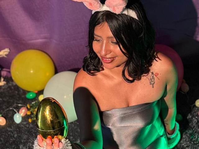 ZarayWhitMoreLove with black wavy hair, pink bunny ears, gray strapless dress, holding a shiny gold Easter egg, smiling, small tattoo on right shoulder, colorful balloons.