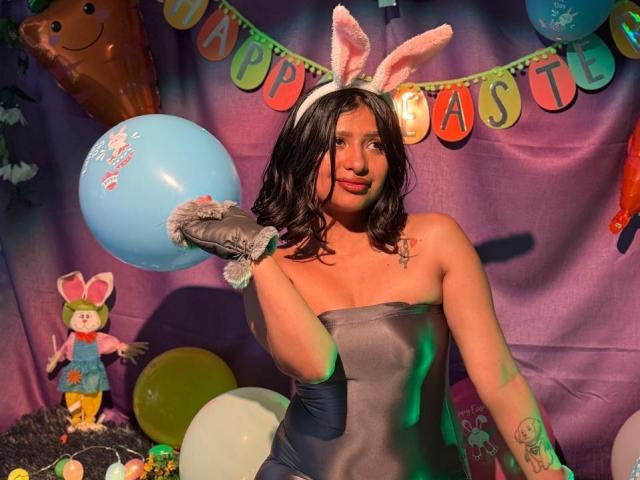 ZarayWhitMoreLove in a grey strapless dress, holding a blue balloon, wearing pink bunny ears and grey gloves, with tattoos on her arm, smiling in a colorful Easter setup.