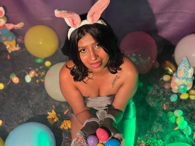 ZarayWhitMoreLove with pink bunny ears, black wavy hair, and gray strapless outfit, holding colorful eggs, tongue out, tattoo on right chest, wearing gray gloves, surrounded by balloons.