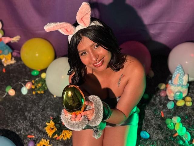 ZarayWhitMoreLove with black hair, pink bunny ears, holding a shiny golden egg, wearing a strapless outfit, smiling, sitting on a gray carpet with colorful balloons and candy.