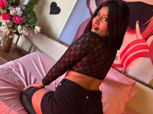 ZarayWhitMoreLove with black lace top, showing her back, black shorts, and black thigh-highs, kneeling on pink bed, looking over her shoulder, with pink roses in the background.