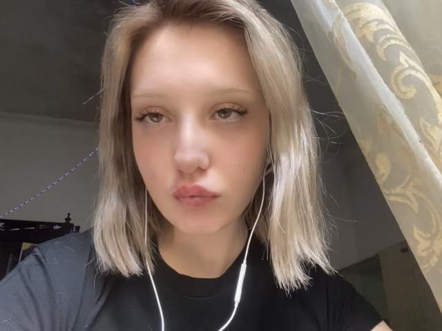 SashaSmithe with blonde hair, black shirt, and white earbuds, pouting with slightly parted lips, looking directly at the camera. Her green eyes are slightly open.