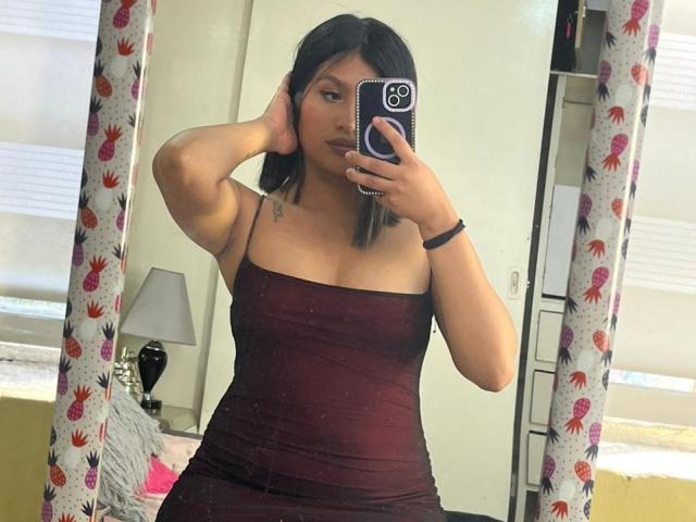 ZarayWhitMoreLove with shoulder-length black hair, wearing a tight, dark red, strapless dress, holding a phone in front of a mirror, hand in hair, visible small tattoo on chest.