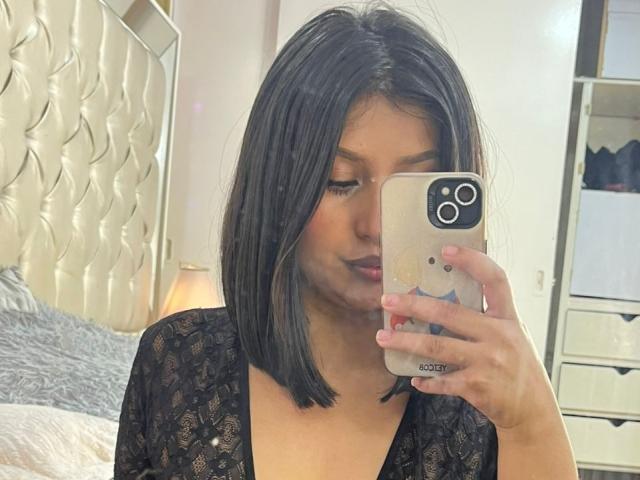 ZarayWhitMoreLove with black lace top, holding phone with bear case, taking selfie. She's got medium tits, straight black bob, pink nails, and is looking down. Bedroom setting.