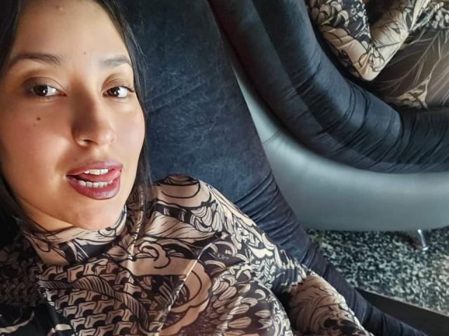 CarmilleZlata with brown eyes, pink glossy lips, wearing a patterned brown and beige top, sitting on a black, textured sofa, looking at the camera with a slight smile.