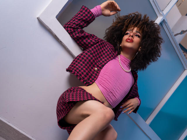 KataleyaFowler with curly afro, wearing pink crop top, houndstooth jacket, and skirt, showing her bare thigh and belly, pearl necklace, red lipstick, and confident pose.