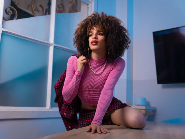 KataleyaFowler with curly afro, pink long-sleeve top, pink patterned shorts, pearl necklace, and red lipstick, kneeling on table, holding jacket, blue walls, window, and flat screen.