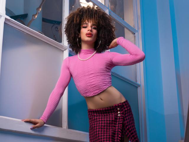 KataleyaFowler with curly brown hair, pink textured crop top, pink and black houndstooth skirt, pearl necklace, standing by window, left arm on ledge, right arm bent.