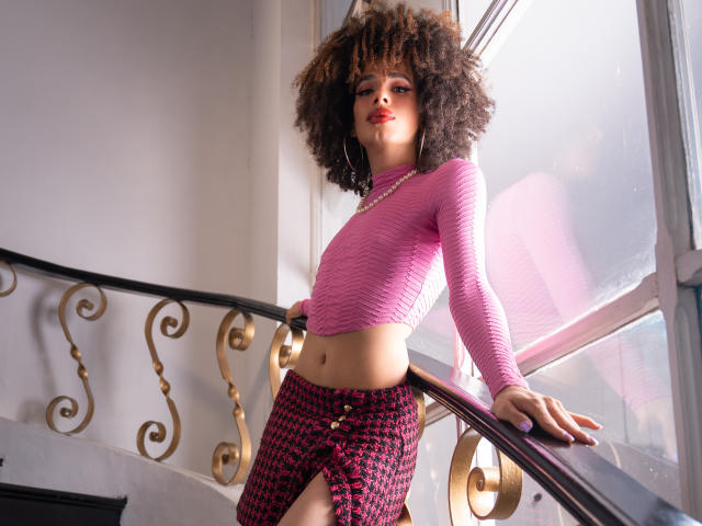 KataleyaFowler with curly brown hair, pink long-sleeve crop top, and pink houndstooth skirt, leaning on a staircase, wearing hoop earrings, standing by a window.