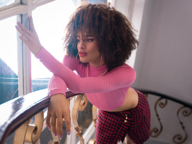 KataleyaFowler with curly brown hair, pink crop top, and red checkered pants, leaning on a wooden railing, sunlight in background, looking to the side with pouty lips.
