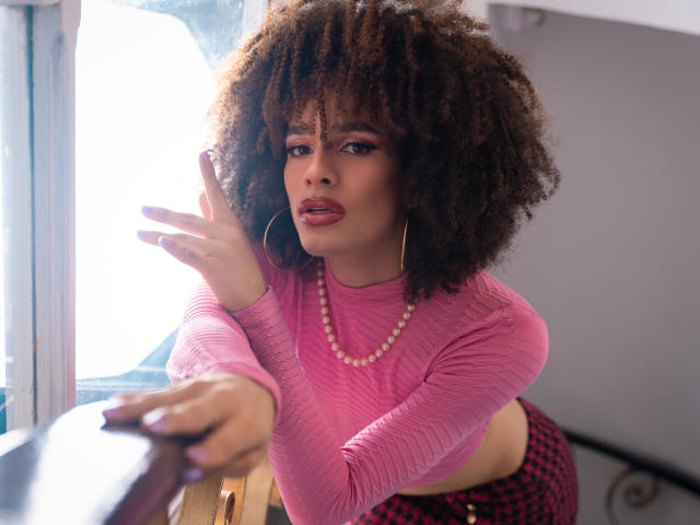 KataleyaFowler with curly brown hair, pink ribbed top, pearl necklace, red checkered pants, leaning on table, hand raised, looking at camera, large hoop earrings, pink lipstick.