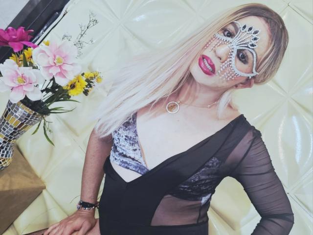 NicolleMorris with white face paint, black sheer dress, showing tits, blonde hair, pink lipstick, and mask, sitting on yellow leather, with flowers in background.