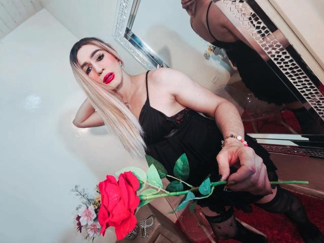 NicolleMorris with long blonde hair, red lipstick, black lace lingerie, holding a red rose, reflected in a mirror. She's wearing black stockings and has a bracelet on her wrist.