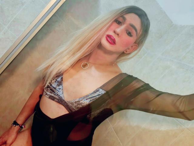 NicolleMorris with long blonde hair, red lipstick, wearing a black sheer top, silver velvet bra, and a gold necklace, taking a selfie in a bathroom. Boobs visible.