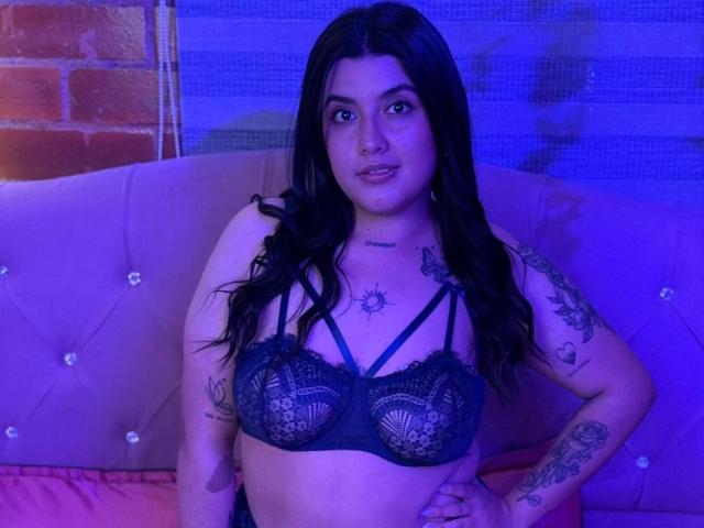 CherrySpicy mit langen, schwarzen Haaren, trägt einen schwarzen Spitzen-BH, zeigt ihre Brüste, hat Tattoos an Armen und Brust, sitzt auf einem pinken, gepolsterten Bett, blaues Licht..