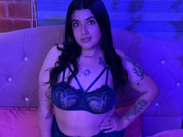 CherrySpicy, avec de longs cheveux noirs, portant un soutien-gorge en dentelle noire qui révèle ses seins, assise sur un canapé rose avec des tatouages sur ses bras et sa poitrine, sous un éclairage bleu..