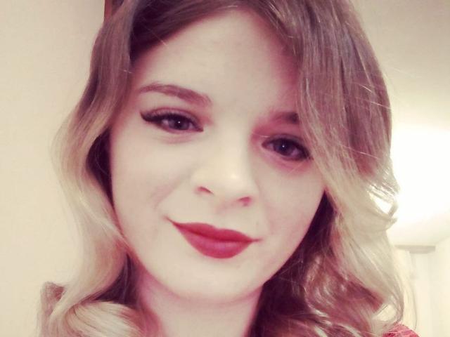 MicheleJayme has wavy blonde hair, red lipstick, and black eyeliner, smiling softly with fair skin and blue eyes, looking cute and sexy.