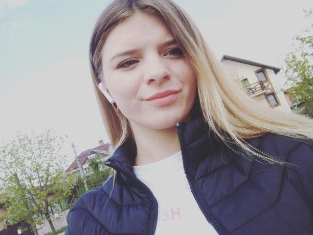 MicheleJayme, avec des cheveux bruns clairs, portant une veste matelassée bleu marine, une chemise blanche et des écouteurs blancs, souriant légèrement, en extérieur, avec des arbres verts en arrière-plan, un ciel bleu, un selfie décontracté..