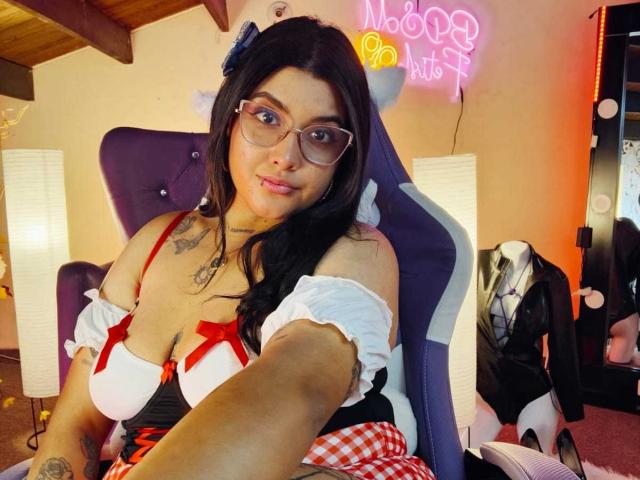 CherrySpicy with glasses, dark hair, and a red-and-white outfit, showing her big tits in a chair, tattoo on her chest, smiling at the camera, wooden ceiling above.