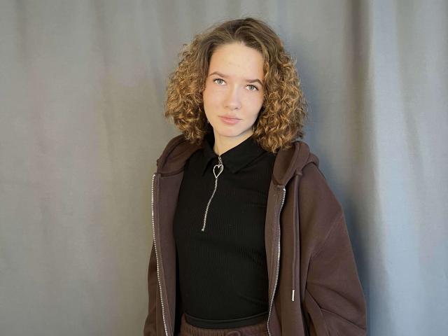 JoellenMasom with curly brown hair, wearing a brown jacket over a black zip-up shirt, standing against a gray curtain, showing a subtle smile. She has small tits and a cute face.