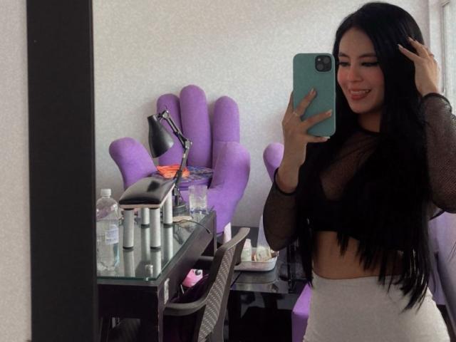EimmyPark with long black hair, wearing a sheer black top, showing her tits, white pants, taking a mirror selfie with a green phone, smiling, in a bedroom.