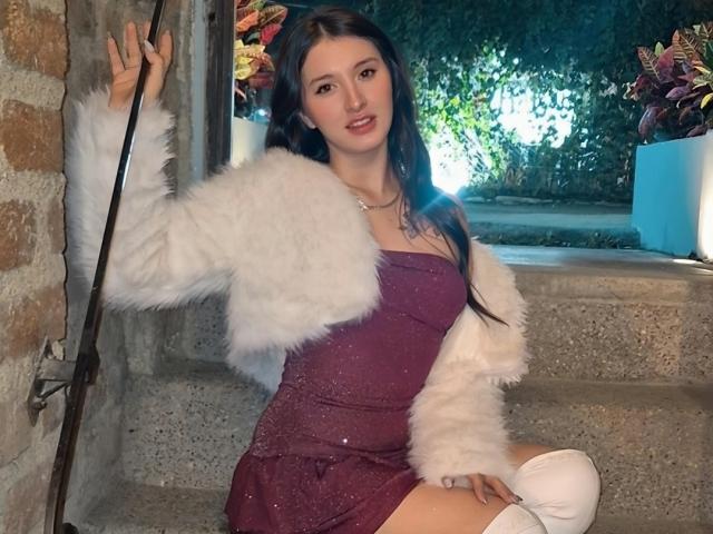 ZoeAvancini with long dark hair, wearing a sparkly purple dress, white fur jacket, and white knee socks, sitting on stone steps, holding a pole with one hand.