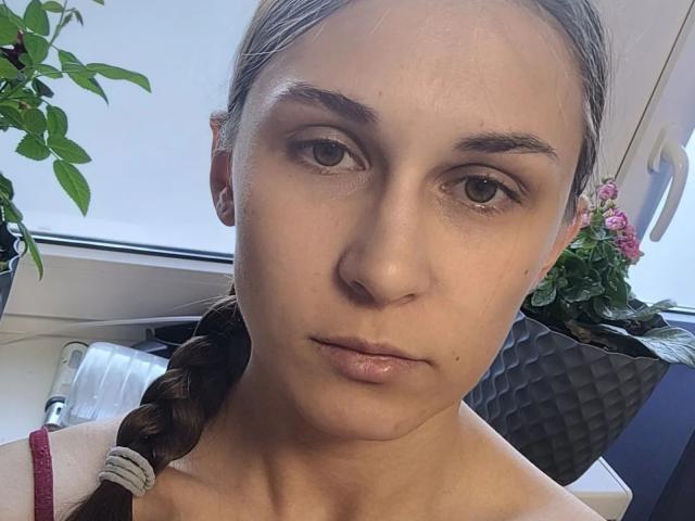 Marie Dunton tiene ojos marrones, una coleta trenzada y labios rosados, con una expresión neutra, lleva una correa roja y pequeños pendientes blancos, en una habitación luminosa..