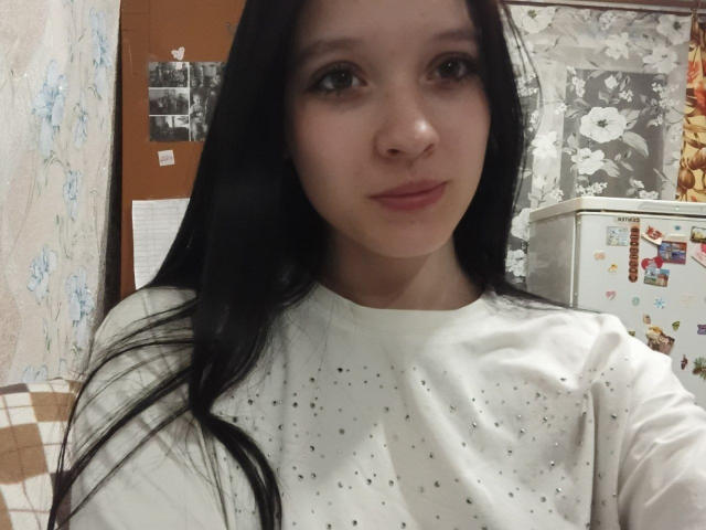 PageMayeux with long black hair, light skin, and small breasts, wearing a white speckled shirt, looking into the camera with a slight smile, in a kitchen setting.
