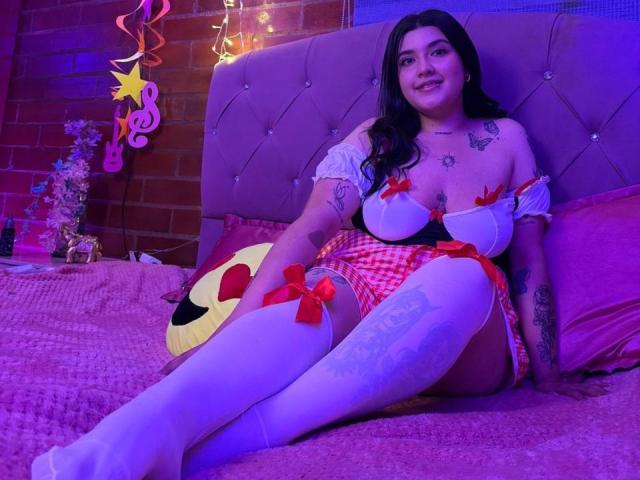 CherrySpicy, com seios grandes e um traseiro curvilíneo, está sentada em uma cama em um quarto iluminado por luzes roxas. Ela está vestindo um conjunto xadrez vermelho com meias altas brancas e laços vermelhos..