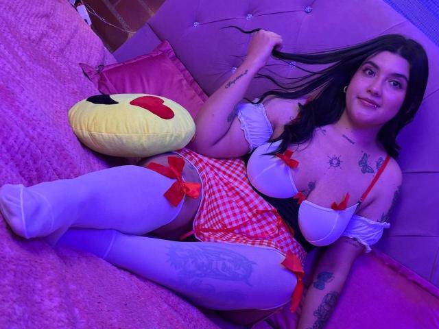 CherrySpicy mit Brüsten in einem weißen BH, einem roten karierten Rock und weißen Oberschenkelstrümpfen, die ihre langen schwarzen Haare hält, mit Tattoos auf Brust und Bein, sitzend auf einem rosa Bett..