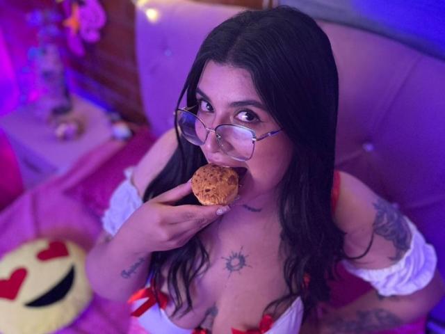 CherrySpicy met een bril, donker haar, tatoeages, die een koekje eet, een witte top met open schouder draagt, een rode strik, die omhoog kijkt, met roze en blauwe verlichting, in een slaapkamer, sexy..