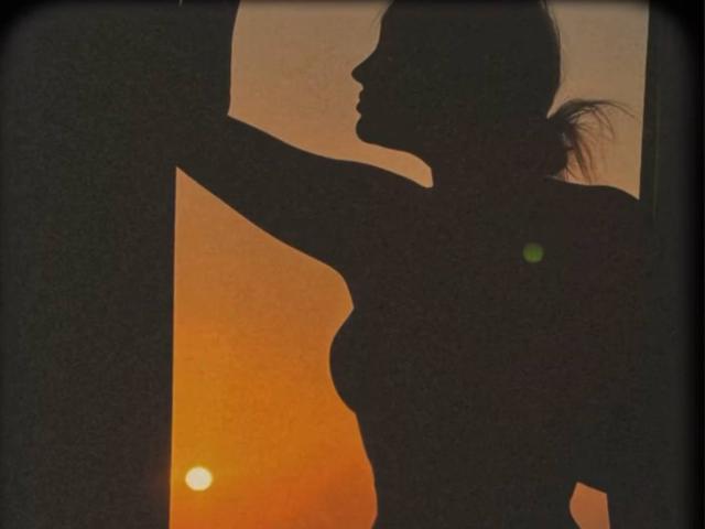 Talitha Gillick, silueteada, con una coleta, levantando el brazo, mostrando pequeños senos, contra un atardecer naranja. Su perfil es oscuro, con un pequeño sol cerca de su parte baja de la espalda..