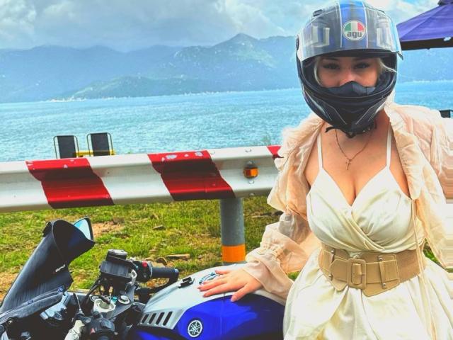 Talitha Gillick mit blauem Motorradhelm, schwarzem Visier, weißem Kleid, beigem Gürtel, sitzend auf einem blauen Motorrad, wobei ihre großen Brüste sichtbar sind, vor einer rot-weiß gestreiften Barriere..