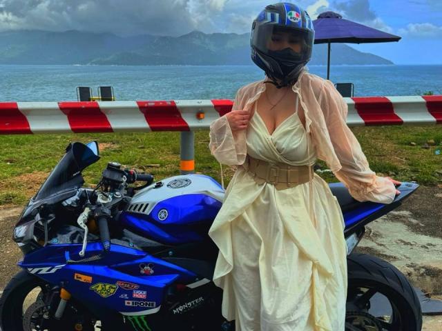 Talitha Gillick, avec de grandes seins, portant une robe beige transparente, un casque bleu et un masque, appuyée sur une moto bleue, au bord de l'eau, avec des montagnes en arrière-plan..