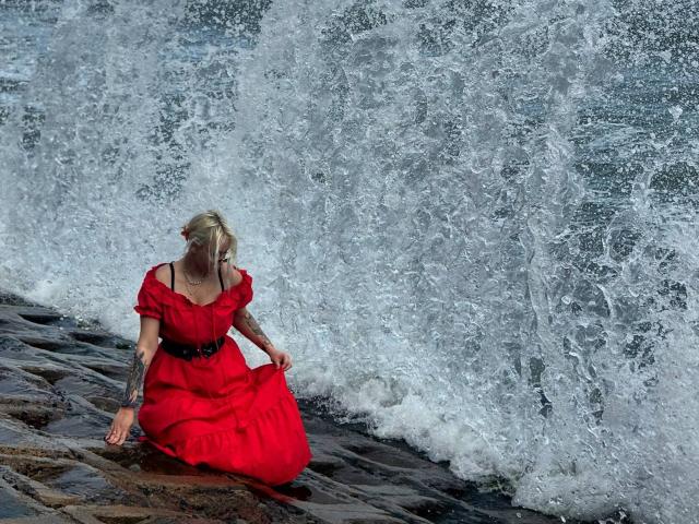 Talitha Gillick, rubia, con un vestido rojo de mangas abullonadas, cinturón negro y tatuajes, arrodillada en una costa rocosa, sosteniendo el vestido, con olas rompiendo detrás de ella..