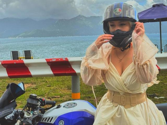 Talitha Gillick indossa un casco trasparente, una maschera nera e un abito beige leggero con una cintura, ed è in piedi accanto a una motocicletta blu, tenendo il casco tra le mani..