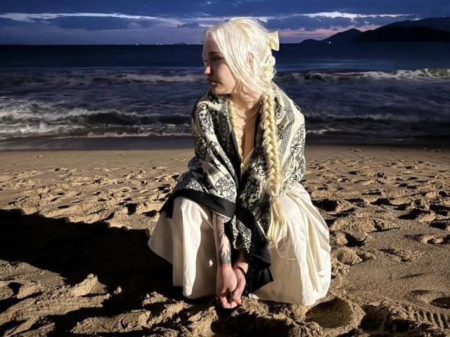 Talitha Gillick tiene el cabello largo, rubio y trenzado, y lleva una blusa negra y blanca con un escote en V pronunciado, así como pantalones blancos, mientras está agachada en una playa arenosa al atardecer..
