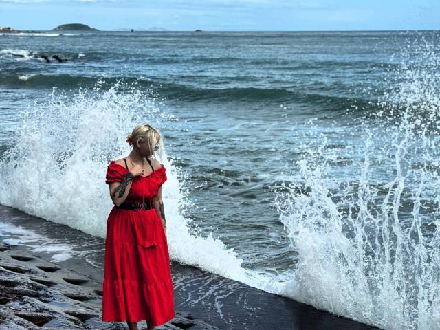 Talitha Gillick è in piedi, indossa un abito rosso, con l'acqua che le schizza intorno, le braccia incrociate e lo sguardo rivolto verso il basso, su una spiaggia rocciosa, con l'oceano sullo sfondo, ha i capelli biondi e dei tatuaggi..