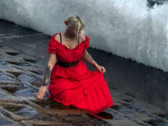 TalithaGillick with blonde hair and a red dress, sitting on rocky shore, waves crashing behind. She's holding dress, tattoos on arms, black belt, and necklace, looking down.