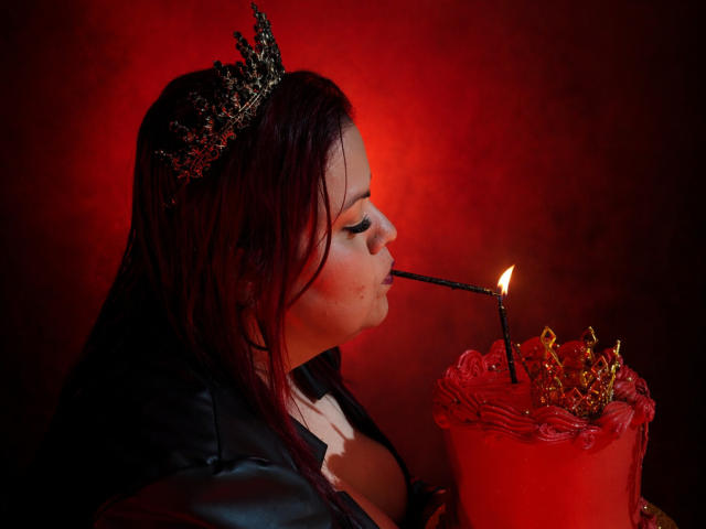 VanesaMoka with dark red hair, wearing a black leather jacket and a glittery crown, sucks a candle flame. Red candle in front, glowing. Her tits are partially visible.