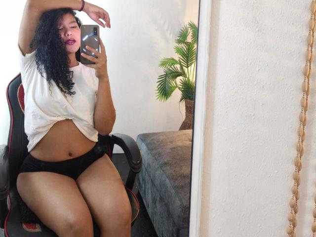 AriaVega with curly black hair, pink lipstick, white tied shirt, black panties, holding phone, arm raised, sitting on black and red chair, reflecting in mirror.