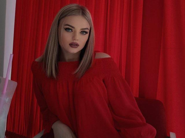 Anisa Healey, met een rode top met open schouder, blond kort haar, donkere make-up en rode lipstick, zittend voor rode gordijnen, kijkend naar de camera, waarbij haar borsten lichtjes zichtbaar zijn onder de top..
