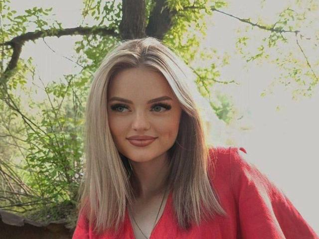 AnisaHealey with blonde, straight hair, wearing a red shirt, smiling with pink lipstick, blue eyes, and subtle makeup, standing outdoors.