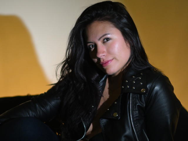 Laura Miller, con lunghi capelli neri, indossa una giacca di pelle nera con borchie, è seduta e guarda la fotocamera con un leggero sorriso. Il suo seno è visibile sotto la giacca..