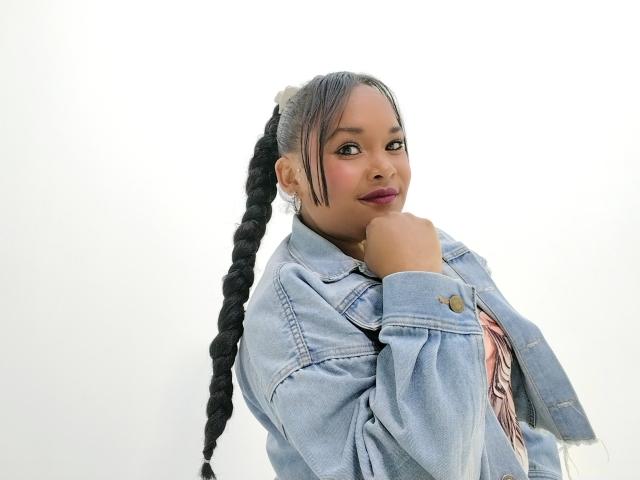 KaylaDark with long black braids, wearing a light blue denim jacket, looking over shoulder with hand near chin, wearing pinkish-red lipstick, standing against a plain white background.