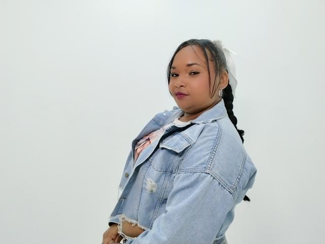 KaylaDark, com cabelo trançado, vestindo uma jaqueta jeans azul clara sobre uma camisa rosa, em pé de lado, olhando para trás com um leve sorriso, mostrando suas nádegas com roupa íntima branca..