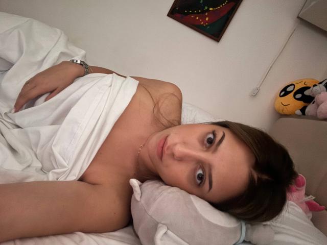 TheaOrre with brown hair, lying in bed, white sheet covering her tits, looking at the camera, wearing a silver watch, resting her head on a pillow.
