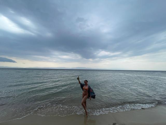 TheaOrre è nuda sulla riva, tiene uno zaino e ha un pene flaccido, circonciso, visibile, sotto un cielo nuvoloso con le onde dell'oceano. Indica a sinistra..